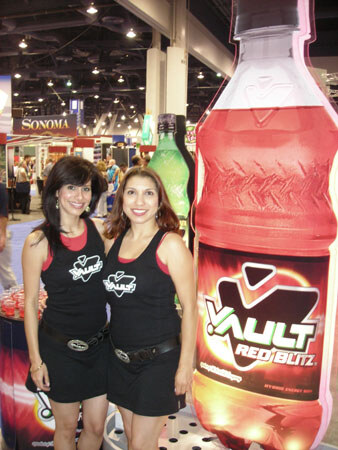 Two smiling women in black Vault outfits stand beside a giant Vault Red Blitz bottle display, with a green bottle and other booths in the hall behind them.