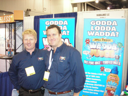 Two men in dark blue shirts with show badges stand together in front of blue Wadda Juice banners reading 'Godda Lodda Wadda?' and 'Spill Proof Wadda Juice'.