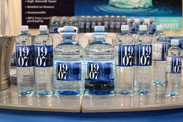Rows of 1907 New Zealand bottled water in small and large bottles with blue labels on a curved counter, with more bottles and a banner in the background.