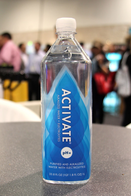A clear bottle of Activate alkalized water with electrolytes and a blue droplet label sits on a gray table, with blurred attendees in the background.