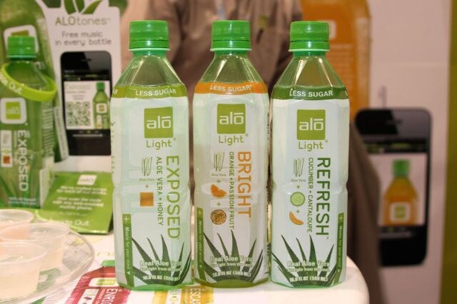Three bottles of Alō Light aloe vera drink: Exposed, Bright and Refresh, in front of an ALOtones sign and sample cups.
