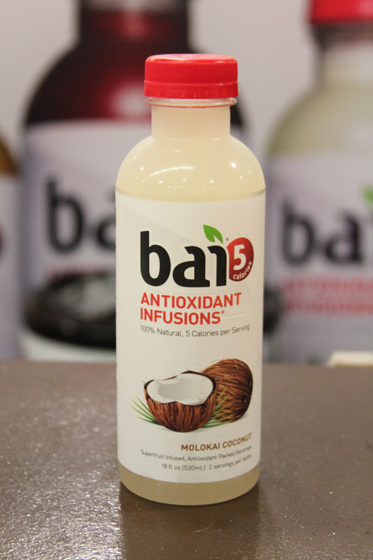 A single bottle of Bai Antioxidant Infusions Molokai Coconut with a red cap on a brown surface, with larger blurred Bai bottles in the background.