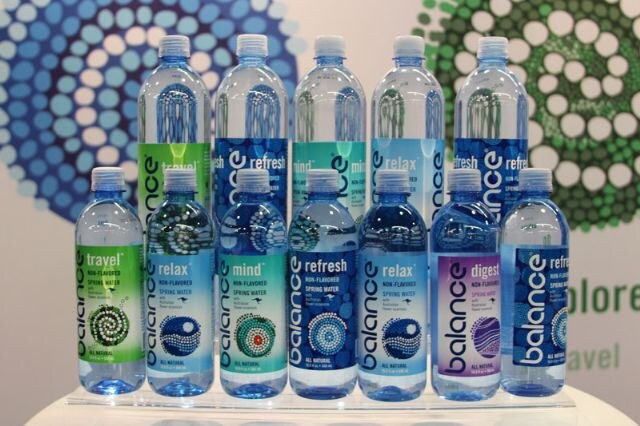 Two tiers of Balance spring water bottles in travel, relax, mind, refresh and digest varieties, arranged on a clear stand in front of blue and green dotted graphics.