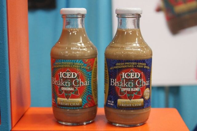 Two glass bottles of Iced Bhakti Chai, Original and Coffee Blend, stand on an orange shelf against a blue and white background.