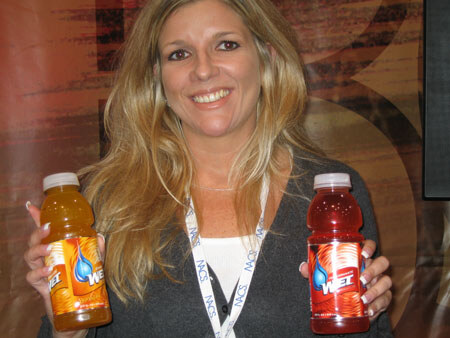 A smiling woman with long blonde hair and a NACS lanyard holds two bottles of WET drink, one orange and one red, in front of a brown backdrop.