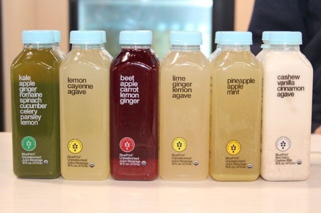 Six BluePrint bottles with light blue caps in a row, holding green, yellow, red, pale and white drinks, with flavor names such as kale apple ginger and cashew vanilla cinnamon agave on the labels.