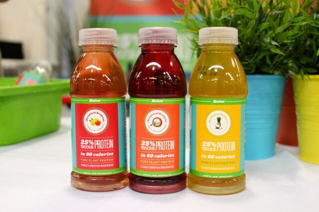 Three bottles of Botan plant protein beverage with red, orange and yellow labels stand on a white table, with potted green plants and a green bin in the background.