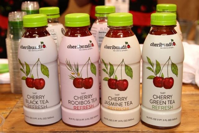 Four bottles of Cheribundi cherry tea drinks with green caps: Black Tea, Rooibos Tea, Jasmine Tea and Green Tea, on a wooden table.