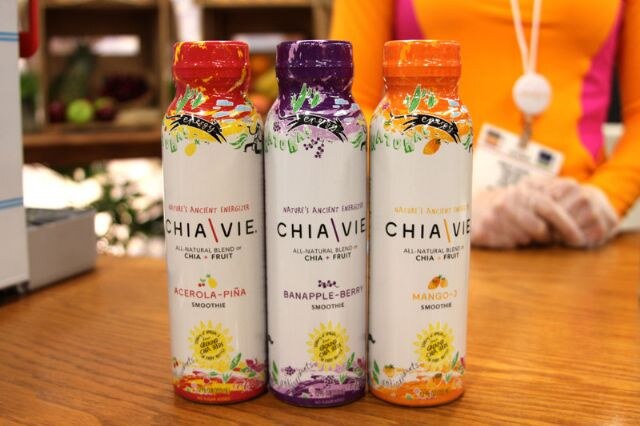 Three bottles of Chia Vie smoothie in acerola-piña, banapple-berry and mango flavors stand on a wooden counter, with a person in an orange top and gloves behind them.