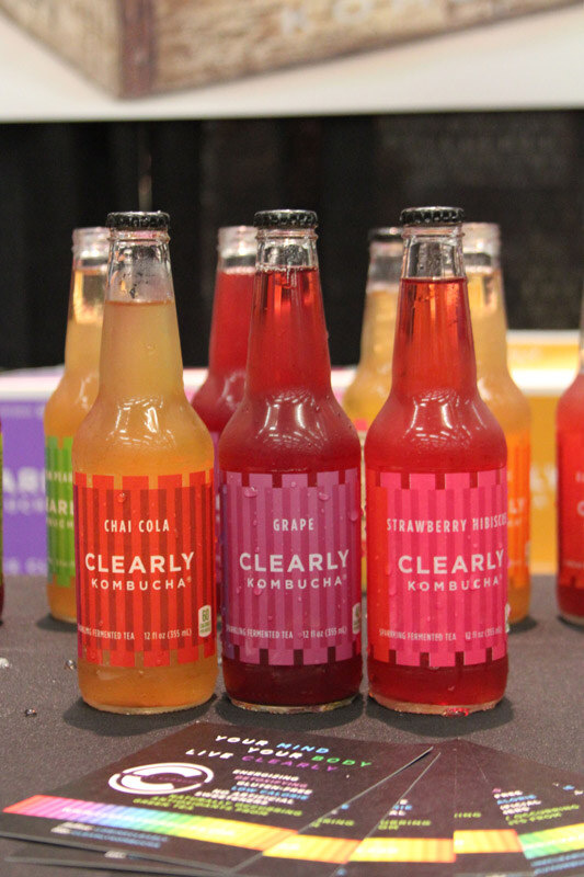 Bottles of Clearly Kombucha in Chai Cola, Grape and Strawberry Hibiscus flavors stand on a dark table, with printed cards in the foreground.