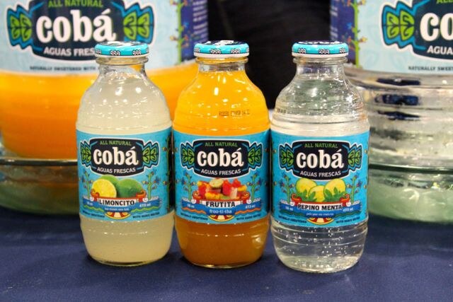 Three Cobá Aguas Frescas bottles in Limoncito, Frutita and Pepino Menta flavors stand on a blue tablecloth in front of larger Cobá containers.