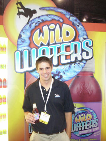 A smiling young man in a black polo and lanyard holds a small bottle in front of a Wild Waters display showing the logo and a large Groovin' Grape bottle.