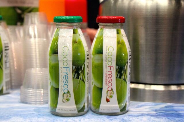 Two small bottles of CocoFresco coconut water, one plain and one strawberry, with green and red caps, in front of stacked clear cups.
