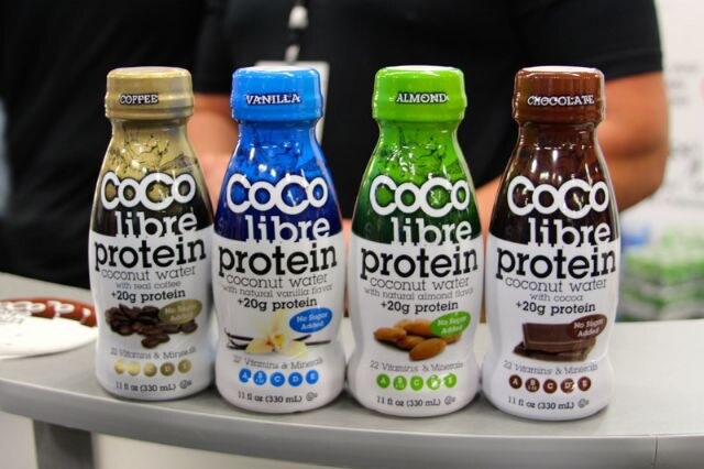 Four Coco Libre Protein coconut water bottles in coffee, vanilla, almond and chocolate flavors stand on a gray counter, with a person in black behind them.