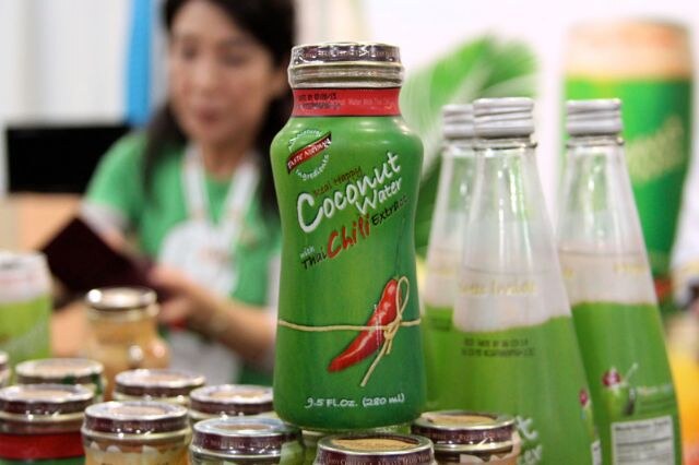 Green bottle of coconut water with Thai chili extract in focus, beside other bottles and jars; a blurred woman in a green top stands in the background.