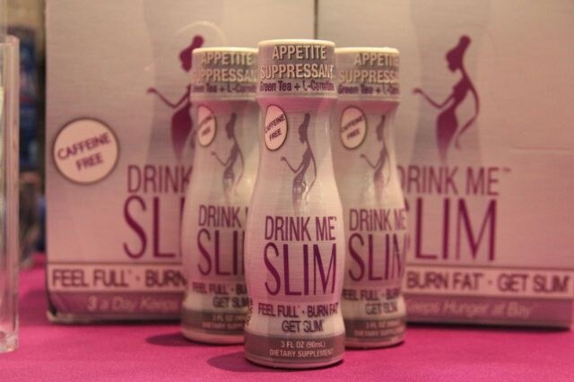 Three small Drink Me Slim appetite suppressant bottles stand in front of a Drink Me Slim box on a pink tablecloth. Labels read Caffeine Free, Feel Full, Burn Fat, Get Slim.