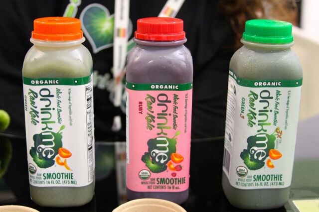 Three drinkme Raw Kale organic smoothie bottles with orange, red and green caps stand on a table, labeled Green, Ruby and Green 3.