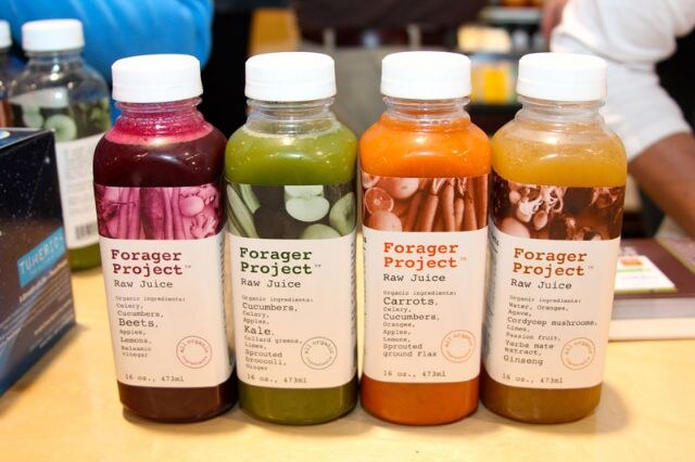 Four bottles of Forager Project raw juice in red, green, orange and amber shades stand on a table, with partly visible people behind them.