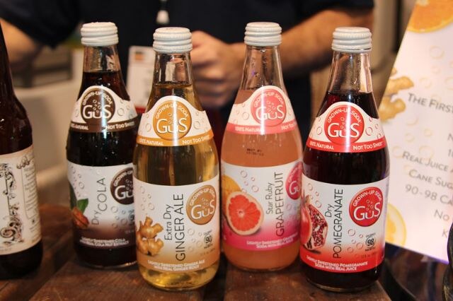 Four bottles of GUS Grown-Up Soda in dry cola, extra dry ginger ale, star ruby grapefruit and dry pomegranate flavors sit on a wooden table, with a person standing behind them.