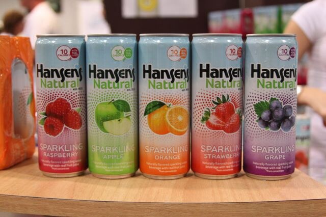 Five Hansen's Natural sparkling juice beverage cans in Raspberry, Apple, Orange, Strawberry and Grape flavors lined up on a wooden counter.