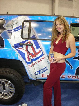 A woman with long wavy hair in a maroon top and pants holds an XL Energy Drink can beside a blue vehicle covered in XL Energy Drink graphics.