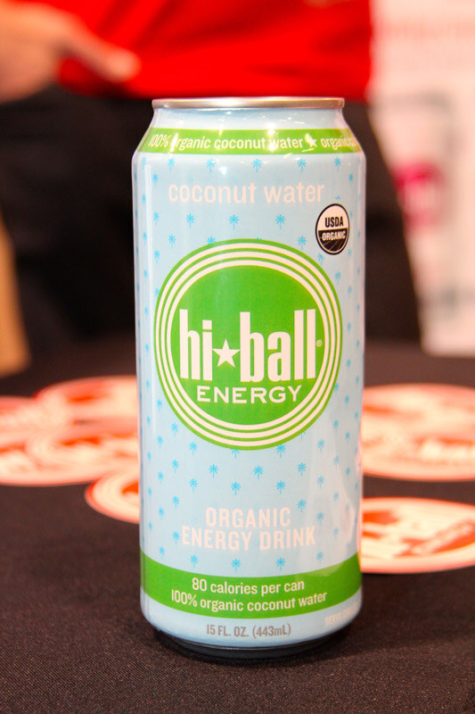 A light blue can of Hi Ball Energy organic coconut water energy drink stands on a black table with round stickers, and a person in red is blurred behind it.