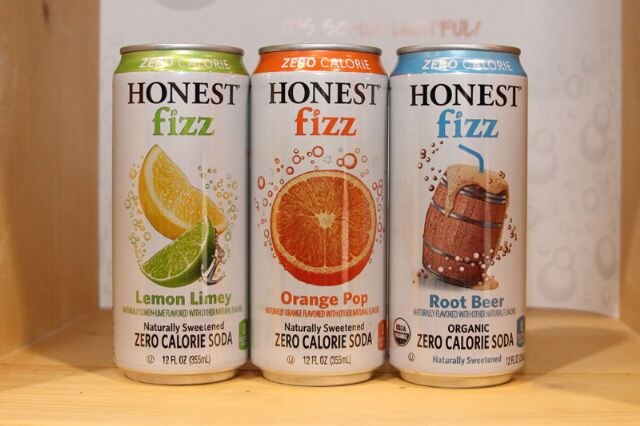 Three cans of Honest Fizz zero calorie soda in Lemon Limey, Orange Pop and Root Beer flavors, standing side by side on a wooden shelf.