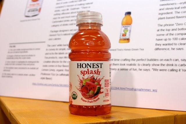 A bottle of Honest Splash Super Fruit Punch with red-orange drink and a watermelon graphic on the label, standing on a wooden shelf in front of printed article pages.