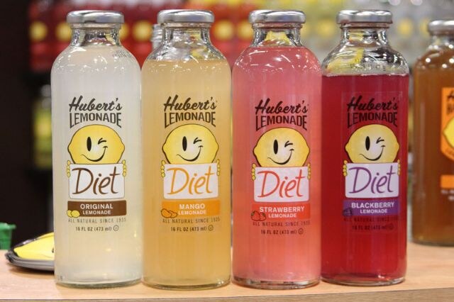 Four bottles of Hubert's Diet Lemonade in a row: Original, Mango, Strawberry and Blackberry, on a wooden counter.
