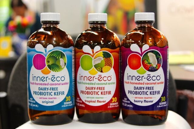 Three amber bottles of inner-eco dairy-free probiotic coconut water kefir, labeled original, tropical flavor and berry flavor, on a chair in front of a blurred background.