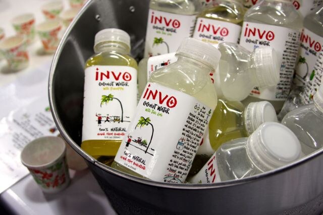 Bottles of Invo coconut water, including pineapple and lime varieties, sit in a metal bucket, with small sample cups nearby.