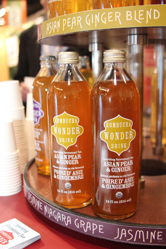 Bottles of Kombucha Wonder Drink Asian Pear and Ginger sit on a round display shelf with flavor names printed on its edge, and a stack of cups at left.
