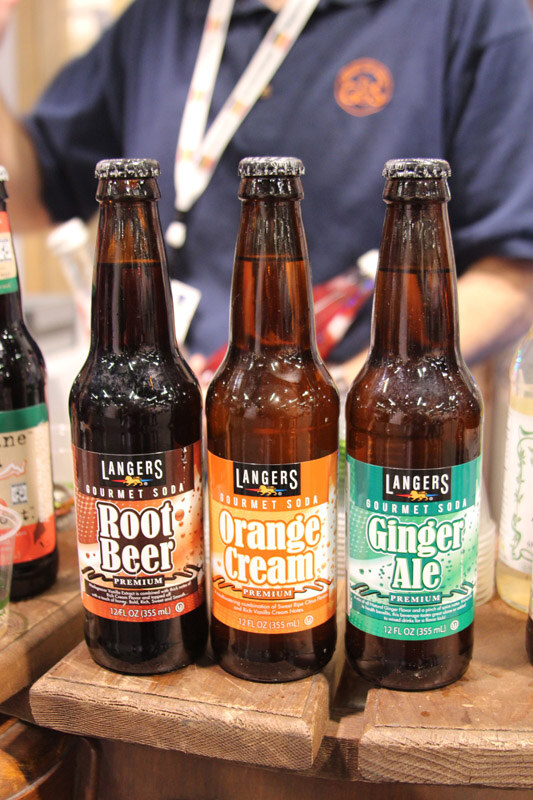 Three brown glass bottles of Langers Gourmet Soda in Root Beer, Orange Cream and Ginger Ale flavors on a wooden surface, with a person in a navy shirt and lanyard behind them.
