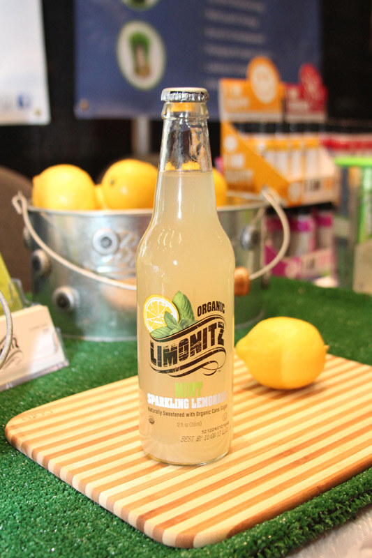 A bottle of Limonitz Organic Mint Sparkling Lemonade stands on a striped cutting board with a lemon, with a bucket of lemons and artificial grass behind it.