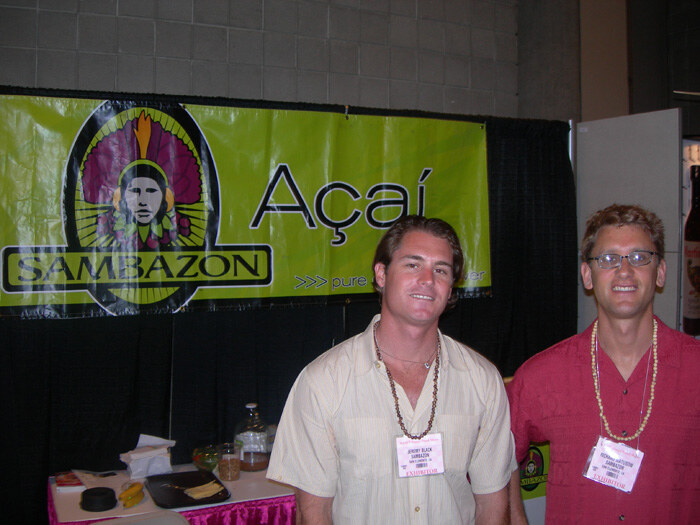 Jeremy Black of Sambazon stands at center in a light shirt and bead necklace, beside a man in a red shirt and glasses, in front of a green Sambazon Açaí banner.