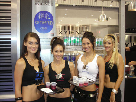 Four women in black and white tops stand at a Xyience booth, two holding trays of small cups, with a Xenergy Clear sign and product cans behind them.