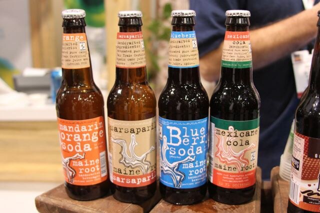 Four glass bottles of Maine Root soda in mandarin orange, sarsaparilla, blueberry and Mexicane cola flavors stand in a row on wooden blocks, with a person partly visible behind them.