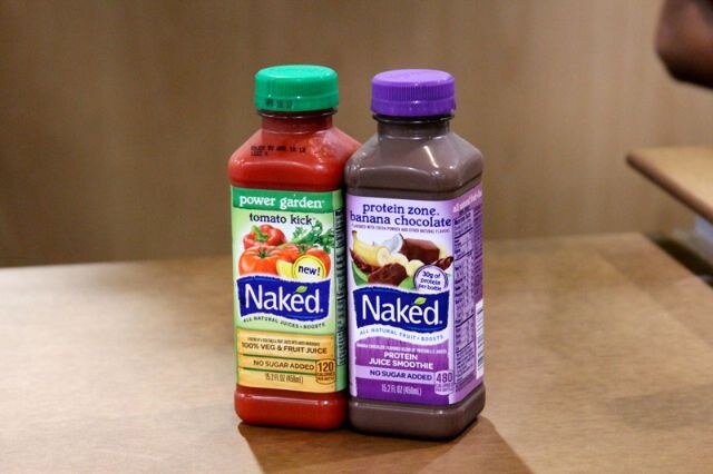 Two Naked juice bottles side by side on a wooden table: Power Garden Tomato Kick with a green cap and Protein Zone Banana Chocolate with a purple cap.