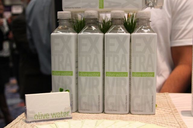 Four bottles labeled Extra Virgin Olive Water from Nurture by Nature on a woven mat, with a card holder in front and a person in a white shirt behind.