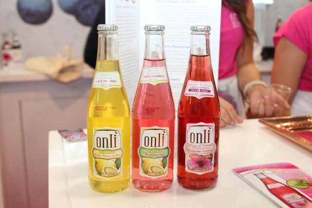 Three Onli glass bottles with yellow, light pink and red drinks on a white table, with two people in pink tops standing behind them.