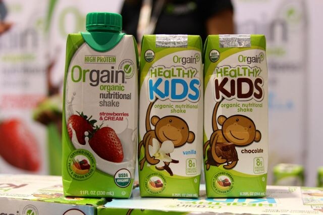 Three Orgain cartons stand on stacked boxes: a strawberries and cream organic nutritional shake and Healthy Kids shakes in vanilla and chocolate, with a person blurred behind.