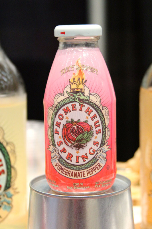 A bottle of pink Prometheus Springs Pomegranate Pepper drink with a dragon and crown label sits on an upturned metal cup, with other bottles blurred beside it.