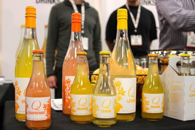 Eight bottles of Q sparkling drinks in grapefruit, orange and lemon flavors stand on a table, with a cardboard four-pack carrier at right and people with badges standing behind.