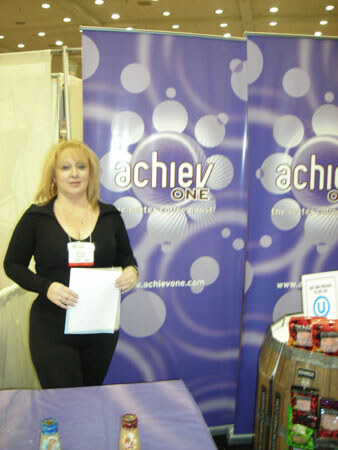 A blonde woman in black holds white papers in front of purple Achiev One banners with bubble patterns. Bottles sit on a table in front of her and packaged products are at right.