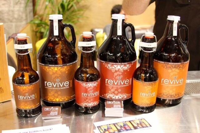 Six amber glass Revive kombucha bottles and growlers in different sizes with brown, red and orange labels stand on a steel counter, with a stack of small cards in front.