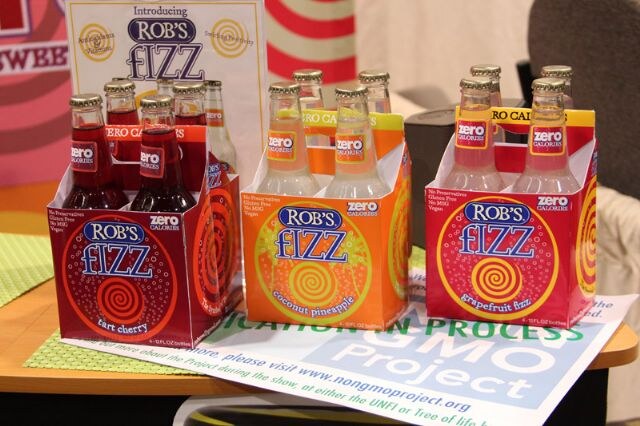 Three four-packs of Rob's Fizz zero calorie soda in tart cherry, coconut pineapple and grapefruit fizz on a table, with a Non GMO Project sign in front.