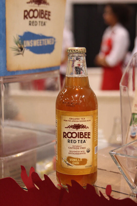 A bottle of Rooibee Red Tea Vanilla Chai sits on a table beside a clear tray, with an Unsweetened Rooibee sign behind it and a blurred person in a red apron in the background.