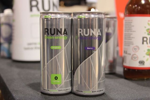 Two silver cans of RUNA clean energy drink, labeled Original and Berry, stand side by side on a dark table, with other RUNA packaging blurred behind them.
