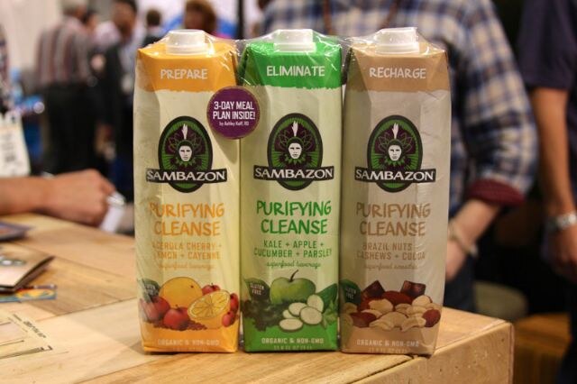Three cartons of Sambazon Purifying Cleanse labeled Prepare, Eliminate and Recharge sit on a wooden counter, with people standing blurred in the background.