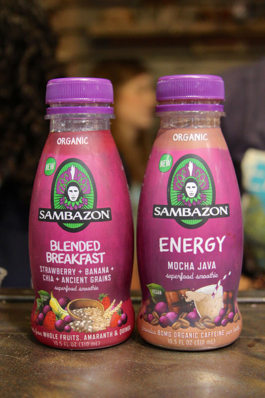 Two purple Sambazon organic superfood smoothie bottles, Blended Breakfast and Energy Mocha Java, stand on a dark surface with a blurred person behind them.